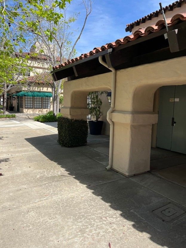 Covered walkway with beige stucco arches beside a courtyard and trees in sunlight