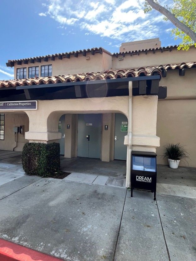 Building entrance with tiled roof, beige stucco walls, and a black signboard on the sidewalk.