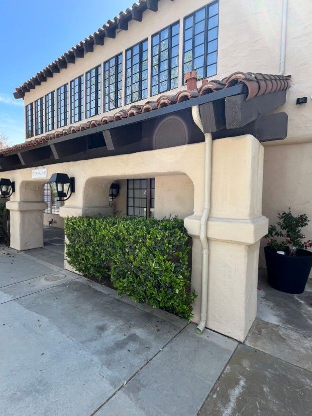 Spanish-style storefront with beige stucco, dark wood trim, large windows, and trimmed hedges along a sidewalk