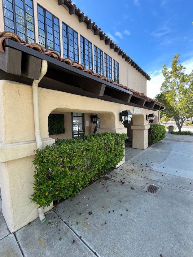 Exterior of a stucco building with arched entry, hedge, and sidewalk under a blue sky