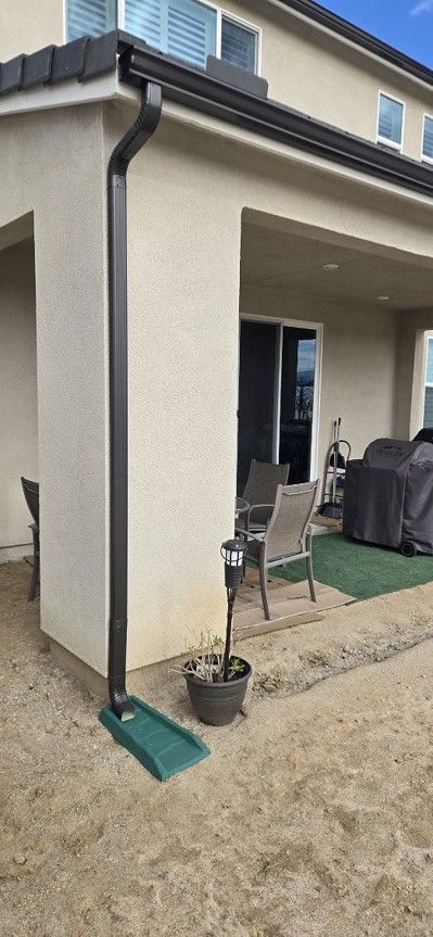 Patio with a beige stucco house wall, sliding glass door, grill, chairs, and small potted plants on gravel.