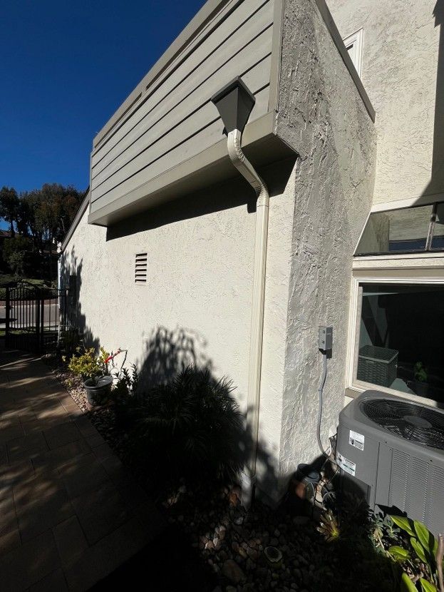 Exterior of a white stucco building with slanted siding, a wall light, and shrubs in sunlight
