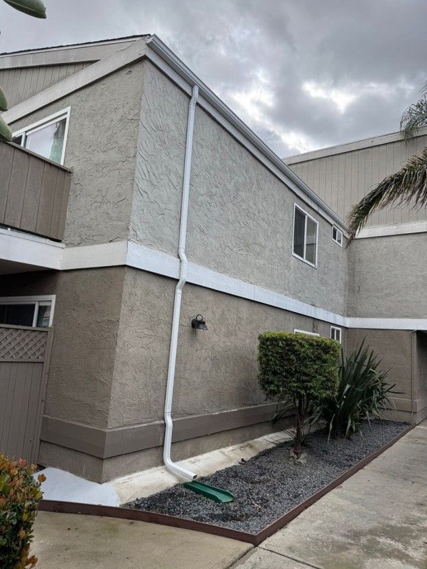 Two-story beige stucco townhouse with white trim, downspout, small shrubs, and a cloudy sky.