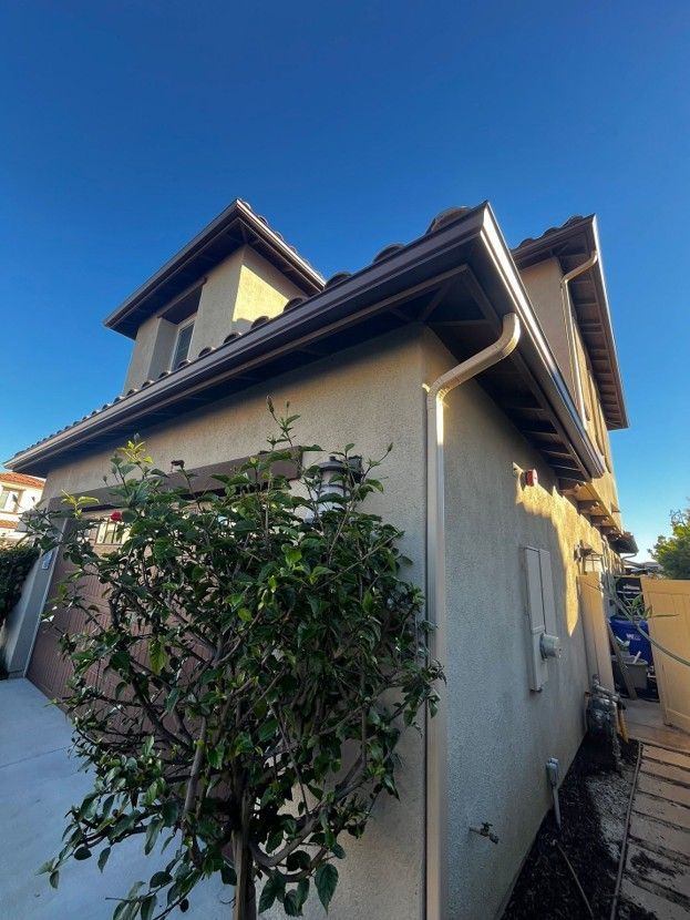 Side view of a beige house with a dark roof, shrubs along the wall, and a narrow walkway beside it