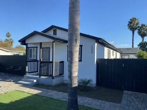 Small white house with black trim, front porch, and a tall palm tree in the yard under a clear blue sky