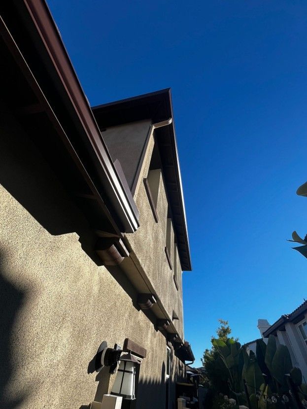Tall beige house wall and eaves under a bright blue sky, viewed from below in a narrow residential alley