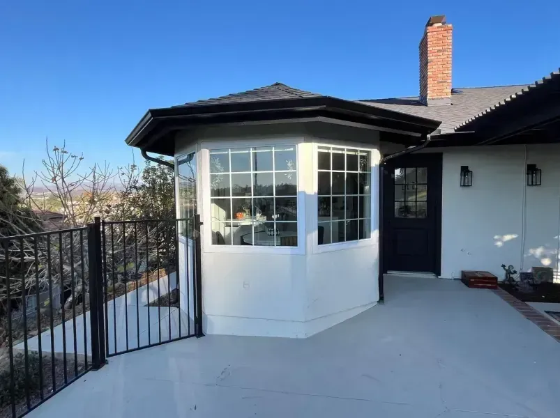 Front porch with white sunroom bay window, black railing, and brick chimney on a sunny house exterior