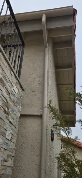 Exterior of a stucco house with a slanted roof, balcony railing, and a small tree below.