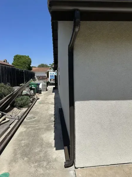 Side yard with concrete walkway, house wall, fence, and a downspout under a clear blue sky
