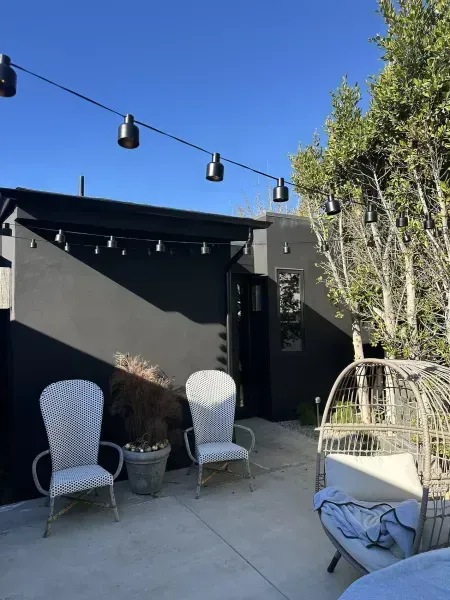 Outdoor patio with two chairs, a hanging swing, string lights, and potted plants beside a black wall.