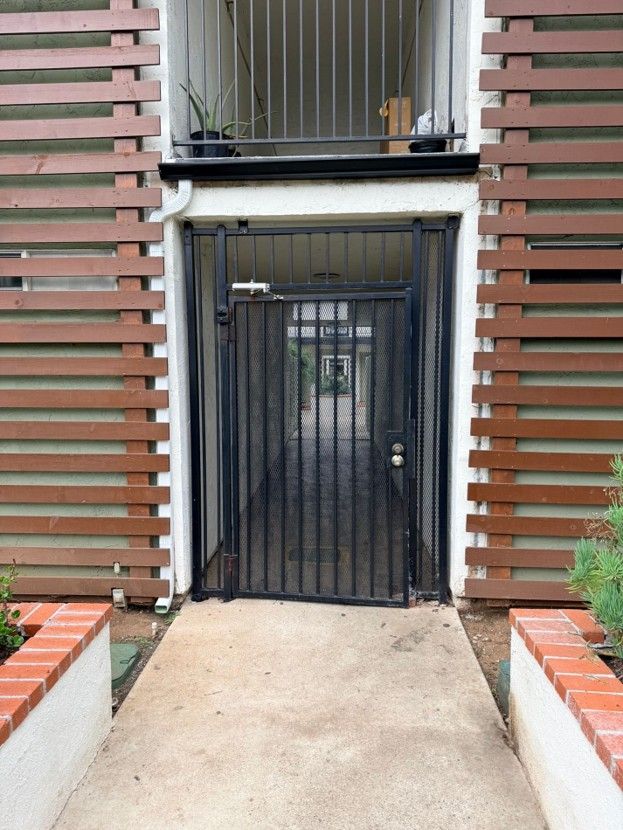 Apartment entrance with a black metal gate between brown slatted walls and a concrete walkway