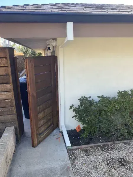 Open wooden gate beside a white house wall and gravel path, with a shrub planter on the right.