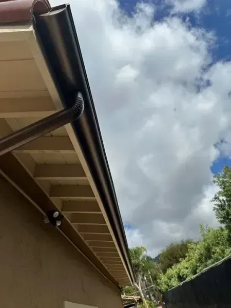 Under a cloudy sky, the underside of a house roof and gutter line above a fence and trees