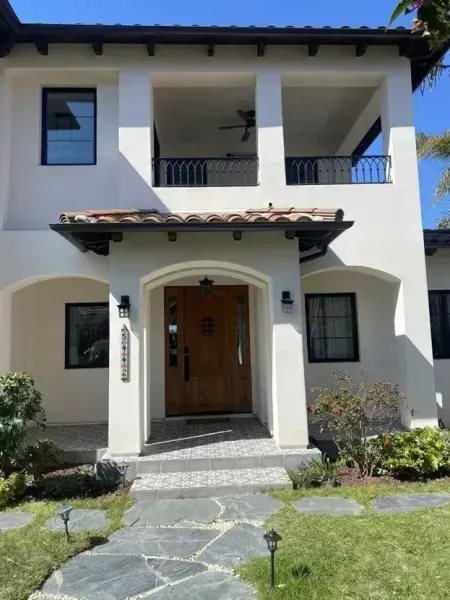 Two-story stucco house with arched entry, wooden front door, and manicured lawn under a clear blue sky