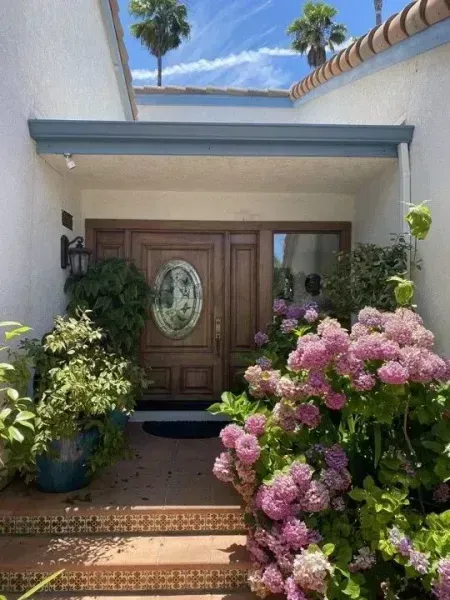 Front entryway with wooden double doors, pink flowers, and potted plants under a blue awning
