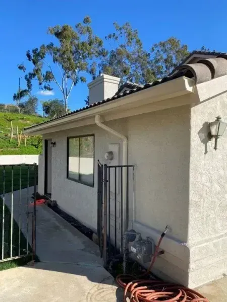 Side view of a beige house with a window, downspout, wall light, and a small fenced patio area