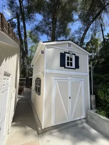 Small white storage shed with black shutters beside a house on a driveway, surrounded by tall trees.