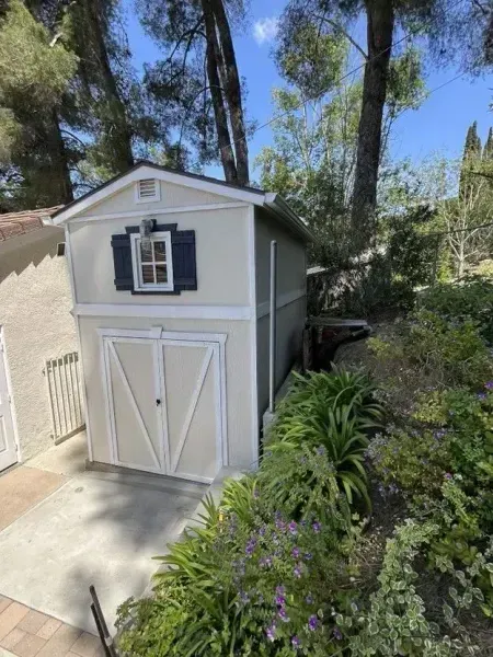Gray backyard shed with white doors and dark shutters beside a concrete path and lush greenery