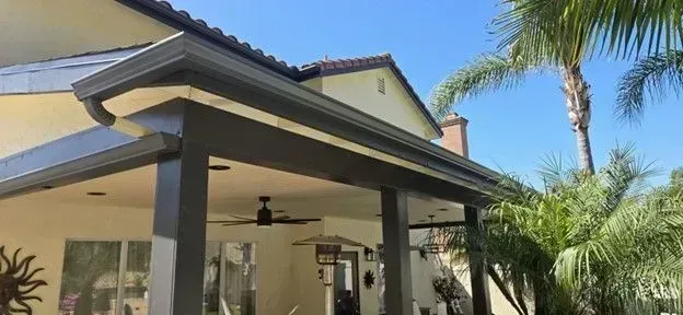 Covered patio with ceiling fans beside a stucco house and palm trees in the yard