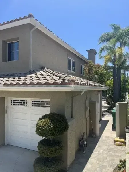 Two-story beige house with white garage, tiled roof, and palm trees beside a narrow walkway.