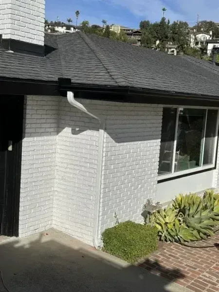 White house exterior with black roof, window, gutter, and a small garden by a brick walkway