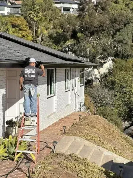 Worker on ladder painting the side of a house beside a steep hillside.