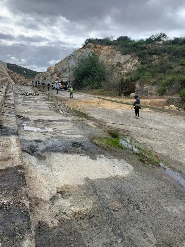Damaged rural road with potholes, small group of people in the distance, and rocky hillside under cloudy sky