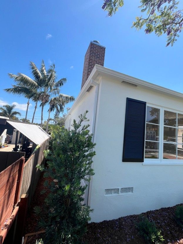 White house exterior with black shutters, brick chimney, palm trees, and a sunny blue sky.