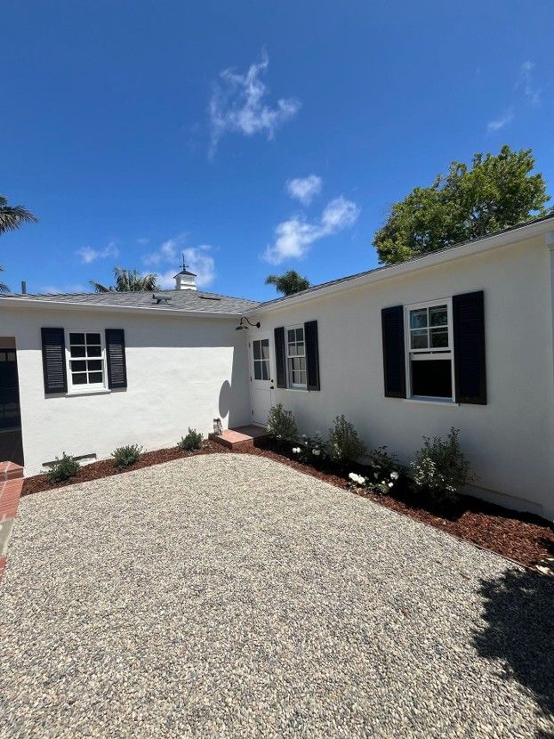 White house exterior with black shutters and a gravel courtyard under a blue sky