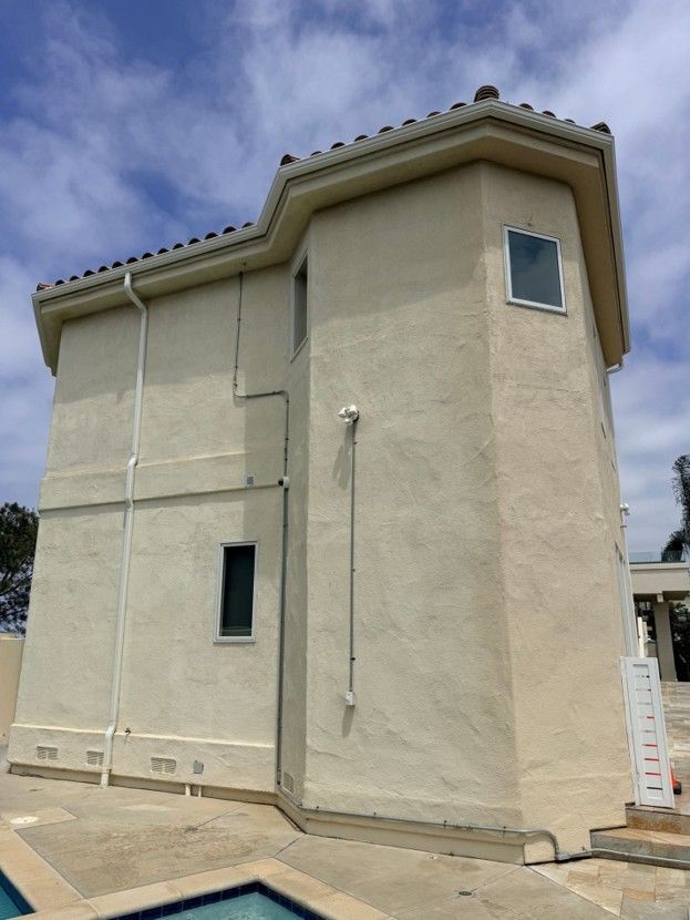 Two-story beige stucco house exterior with a small window and sloped roof under a cloudy sky