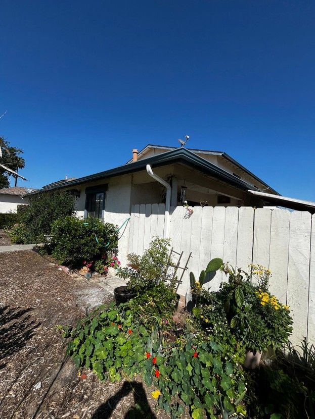 Single-story house with white fence, green shrubs, and a bright blue sky.