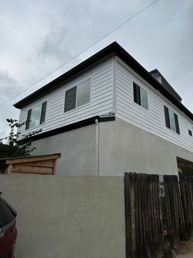 Two-story white house with black trim behind a gray wall and wooden fence under a cloudy sky