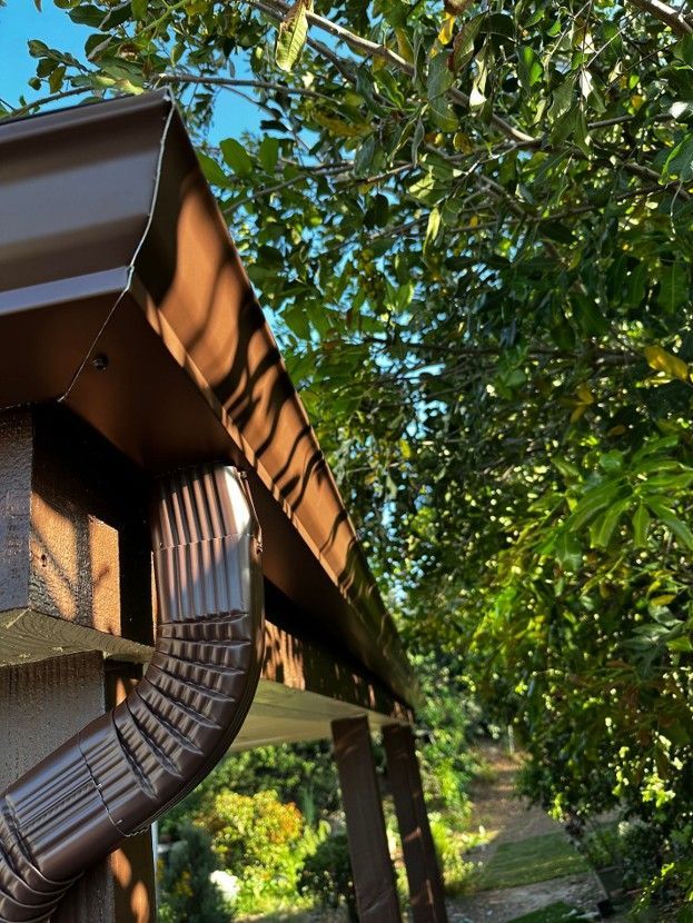 Gutter and downspout on a house roof beside leafy green trees along a sunlit path