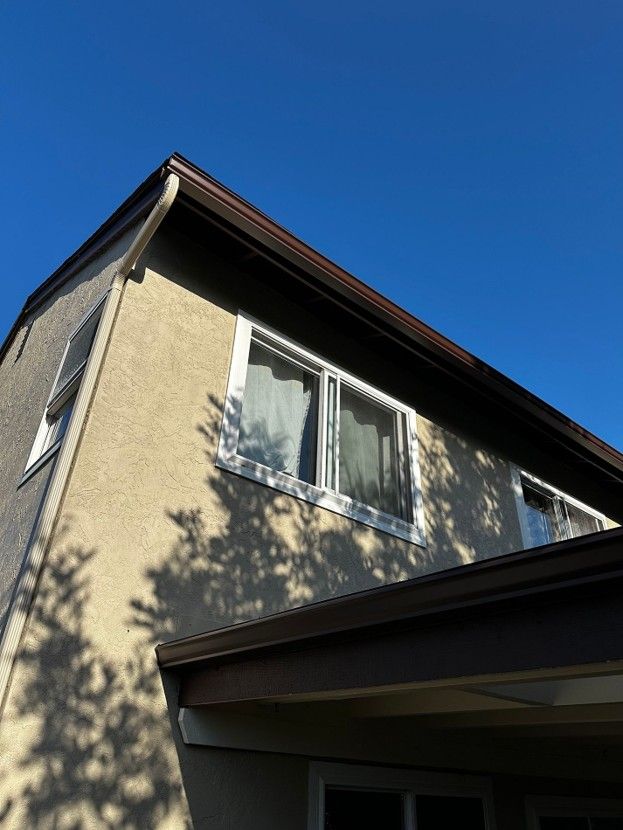 Two-story beige house with a dark roof and a second-floor window against a clear blue sky