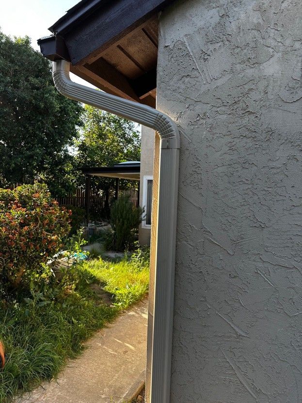 White stucco house wall with a gray downspout beside a garden path and greenery