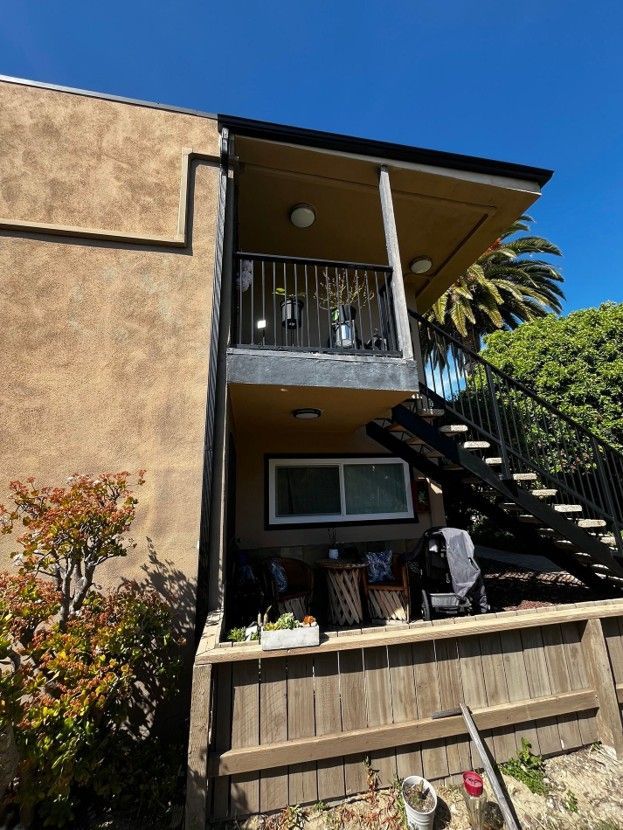 Apartment exterior with tan stucco, balcony, and black staircase beside a fence and greenery