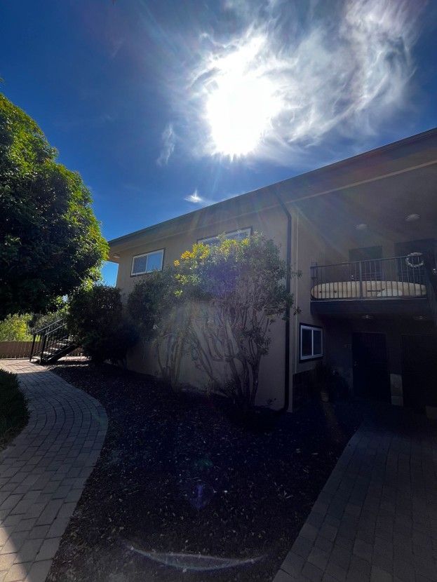 Sunlit apartment building exterior with a balcony, trees, and a curved walkway beneath a bright sky