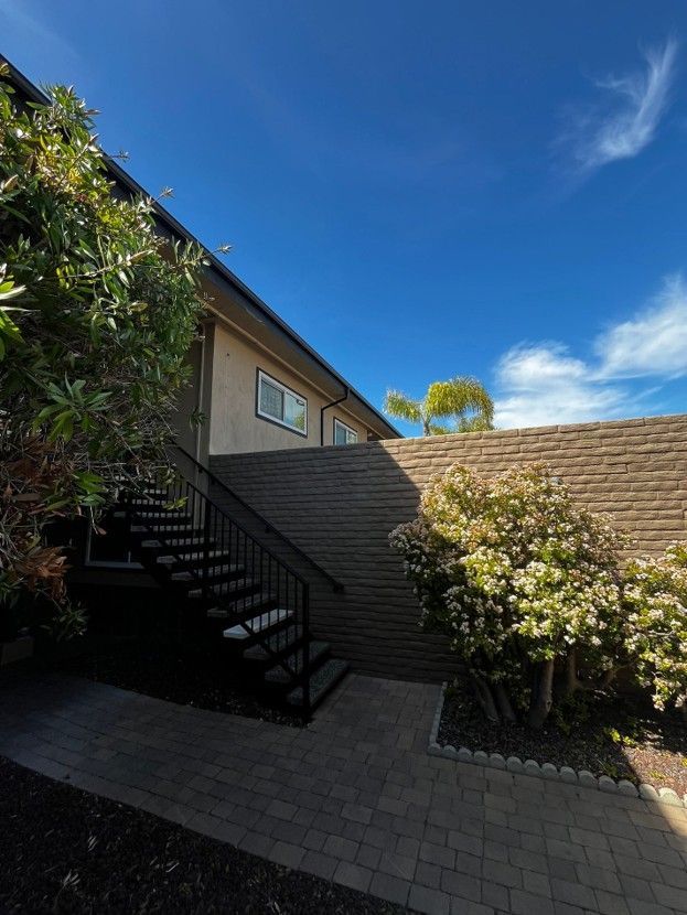Sunlit courtyard with stairs, tan brick walls, green shrubs, and a bright blue sky