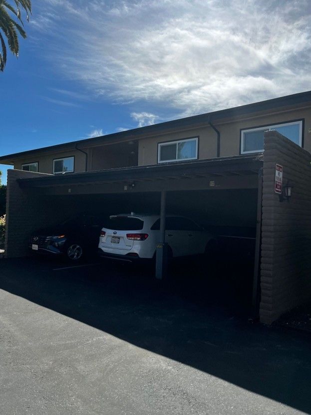 Apartment building with a two-car carport, including a white SUV and a dark car under a blue sky.