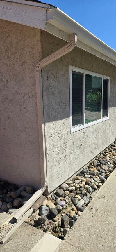 Exterior stucco wall with a window, roof eaves, and rock landscaping beside a concrete walkway
