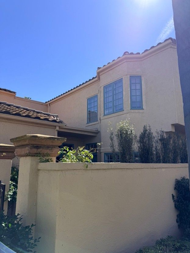 Sunlit beige stucco house with blue-trimmed windows behind a low wall and shrubs