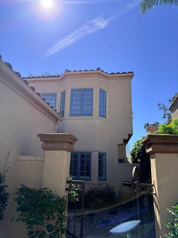 Sunlit beige stucco house with blue shutters and arched windows, viewed from a shaded courtyard.