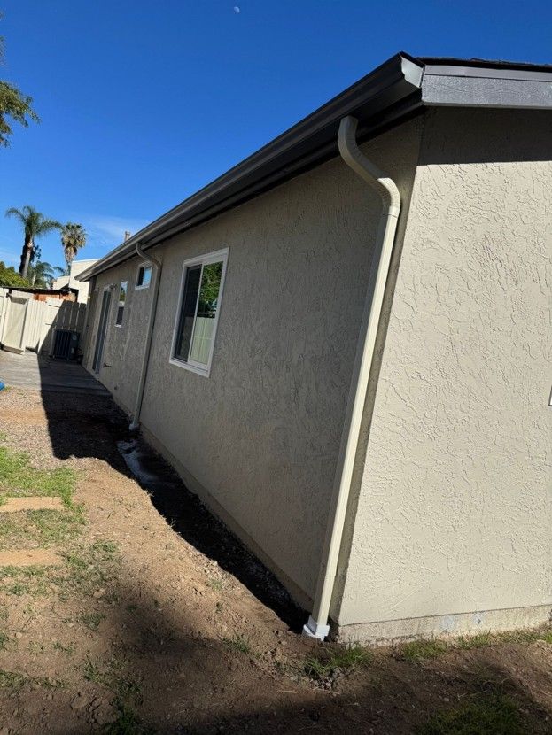 Side view of a beige stucco house with windows, dark roof trim, and a dirt yard under a blue sky.
