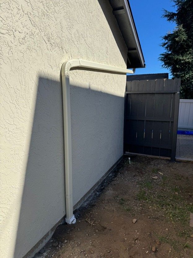 White downspout and horizontal pipe along a stucco wall beside a fenced yard under a blue sky