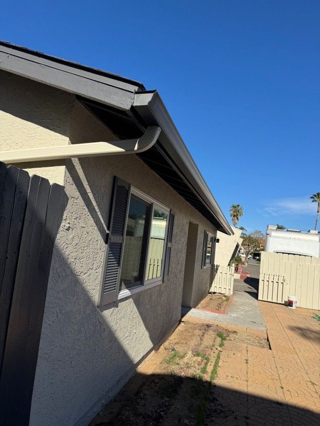 Side view of a gray house with a window, brick path, and beige fence under a clear blue sky