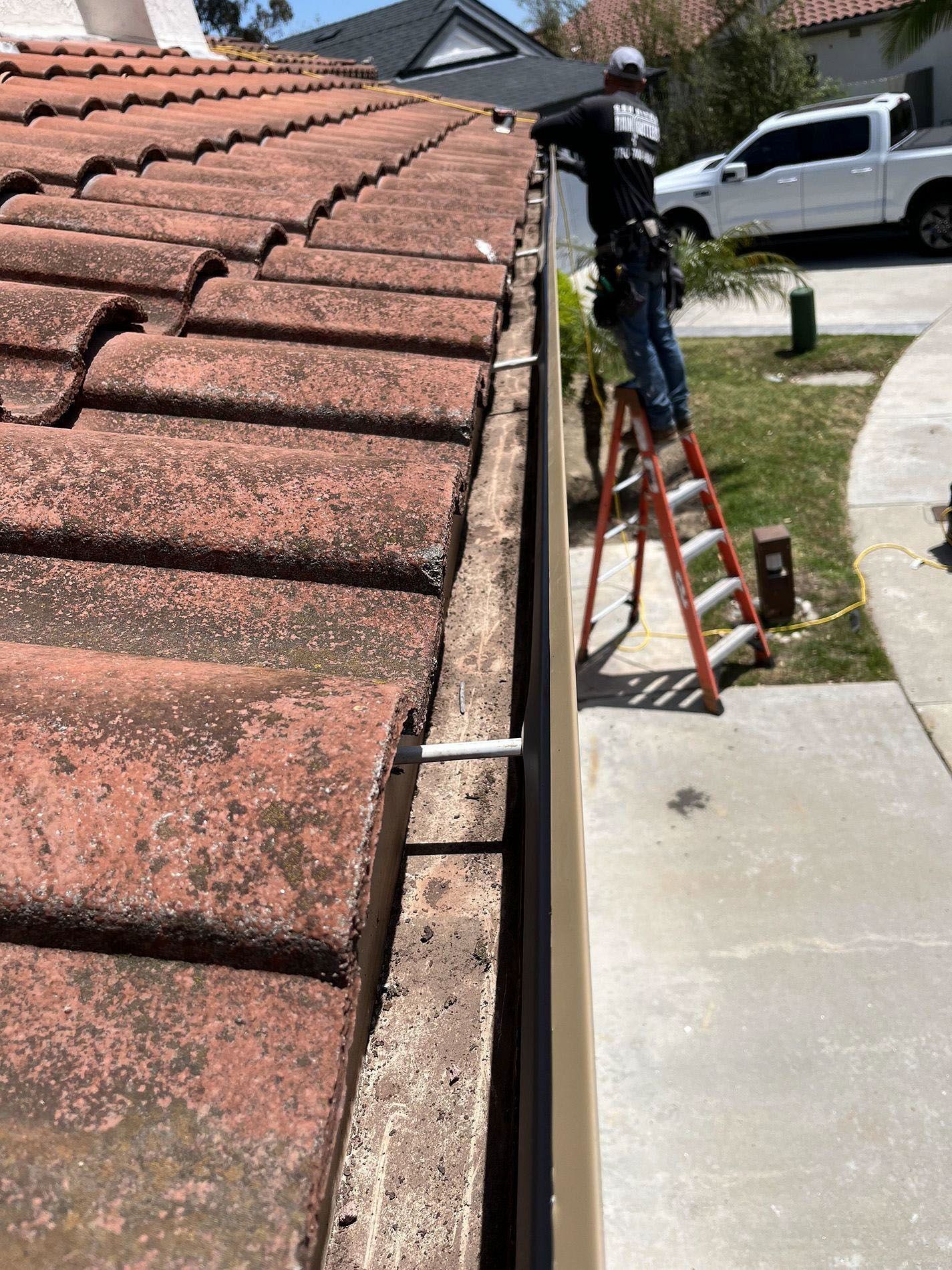 Worker on a ladder repairing the edge of a tiled roof beside a sidewalk and driveway.