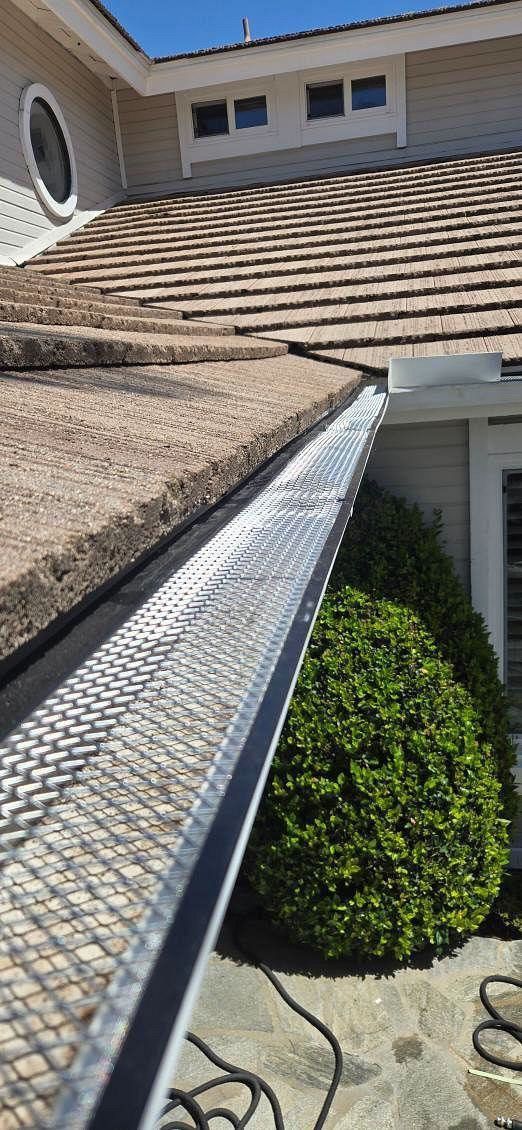 Roof with dark shingles and a ladder-like gutter guard beside a house and green shrubbery