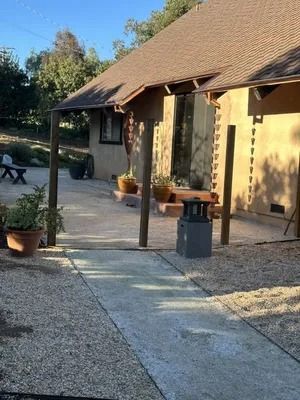 Sunlit patio beside a yellow house with potted plants, stone path, and a picnic table in the background
