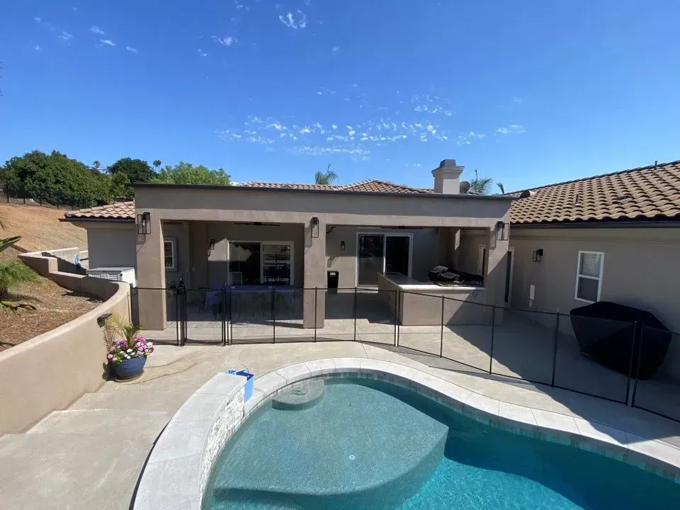 Backyard patio with a swimming pool, covered seating area, and single-story house under a blue sky