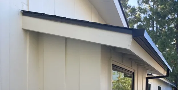 Tan beige house exterior with dark trim and a sloped roof under trees.
